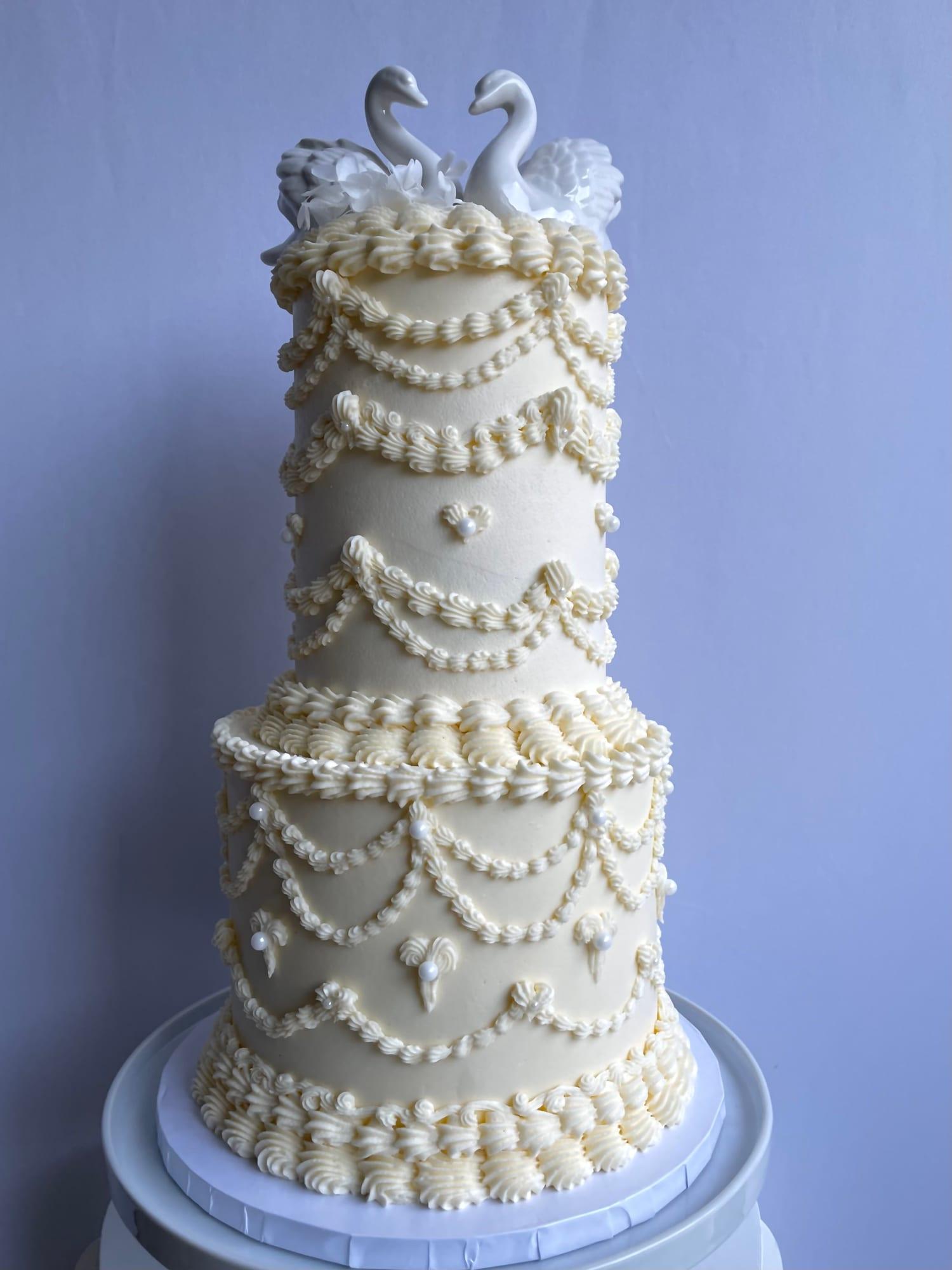 Two-tier ivory wedding cake with Lambeth-style piped swags and pearl beading, topped with porcelain swans